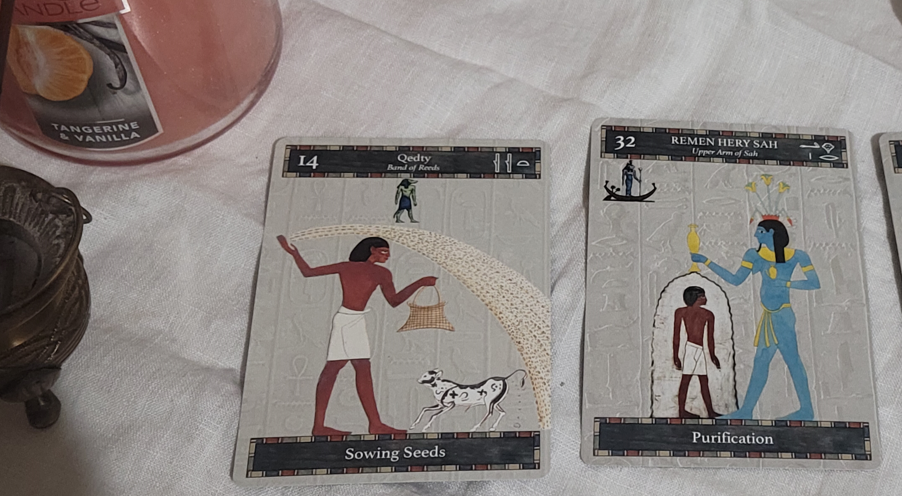 A photo of two oracle cards with Egyptian figures. The one on the left has the figure of a man strewing seeds in front of a cow, and the one on the right has the figure of a man having water poured upon him by the blue-skinned god Hapi.